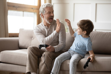 Happy senior grandparent give high five to cute little grandson playing computer games together,...