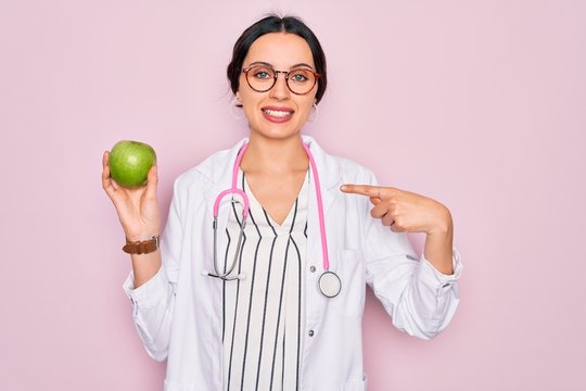 Young beautiful doctor woman with blue eyes wearing stethoscope holding green apple fruit with surprise face pointing finger to himself