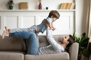 Happy young Caucasian father lying on couch in living room playing with excited little preschooler...