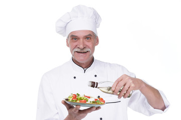 Old male cook holding a plate with vegetables salad and oil on white background