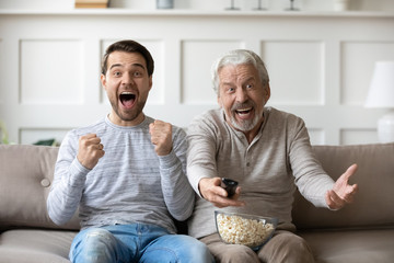 Senior father sit relax on couch with grown-up son watching sports game together, happy mature dad rest at home in living room with adult man child enjoying football match relaxing on weekend