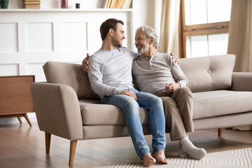 Happy elderly father relax on couch at home with adult son enjoy family weekend together, smiling...
