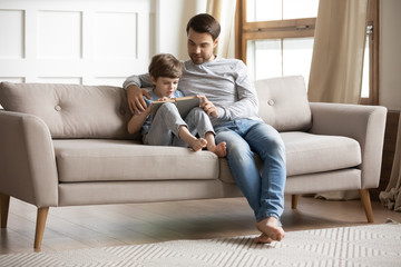 Loving young father and smart little preschooler son sit on sofa in living room learn reading together, caring dad and small boy child relax on couch at home enjoy school interesting book or story