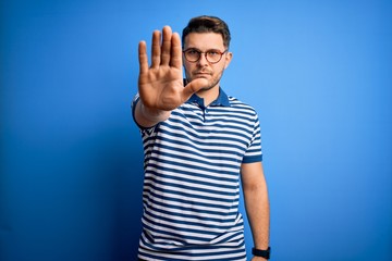 Naklejka premium Young man with blue eyes wearing glasses and casual striped t-shirt over blue background doing stop sing with palm of the hand. Warning expression with negative and serious gesture on the face.