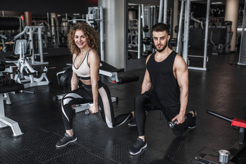Sport, fitness, teamwork, bodybuilding and people concept, young woman and personal trainer with dumbbell squats in gym