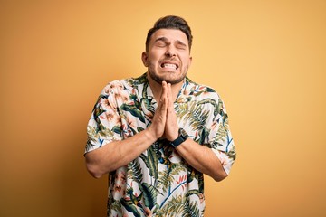 Young man with blue eyes on vacation wearing floral summer shirt over yellow background begging and...