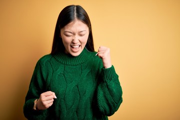 Young beautiful asian woman wearing green winter sweater over yellow isolated background celebrating surprised and amazed for success with arms raised and eyes closed. Winner concept.