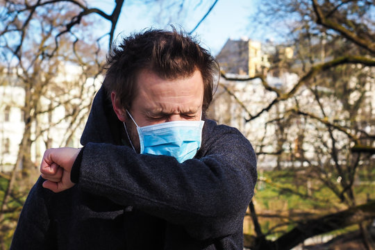 Handsome young European man coughing on the street with a medical face mask on. Closeup of a 35-year-old male in a respirator to protect against infection with influenza or coronavirus (Covid-19) - Powered by Adobe