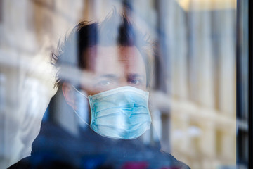 Handsome European man stands behind glass in quarantine with a medical face mask on. Closeup of a 35-year-old male in a respirator to protect against infection with coronavirus (Covid-19). Defocused