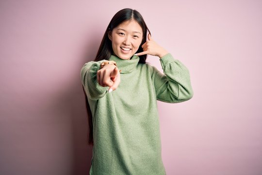 Young beautiful asian woman wearing green winter sweater over pink solated background smiling doing talking on the telephone gesture and pointing to you. Call me.