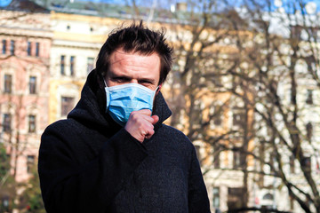 Handsome young European man coughing on the street with a medical face mask on. Closeup of a 35-year-old male in a respirator to protect against infection with influenza or coronavirus (Covid-19)