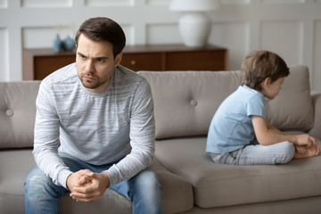 Angry upset young father and little preschooler son sit separate on couch avoid talking after...