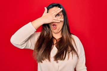 Young hispanic smart woman wearing glasses standing over red isolated background peeking in shock covering face and eyes with hand, looking through fingers with embarrassed expression.