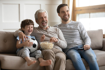 Overjoyed three generations of men relax on couch in living room watch football match eating popcorn together, happy little boy with dad and senior grandfather rest at home enjoy TV game on weekend