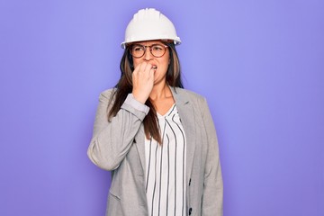 Professional woman engineer wearing industrial safety helmet over pruple background looking stressed and nervous with hands on mouth biting nails. Anxiety problem.