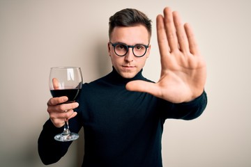 Young handsome caucasian man drinking an alcoholic glass of red wine over isolated background with open hand doing stop sign with serious and confident expression, defense gesture