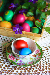 Easter colorful eggs, flowers and greens, white tea-set with eggs inside the cups with butterflies thin porcelain, on a white rounded knitted tablecloth. Country cozy style - happy holiday!