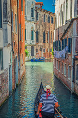 Fototapeta na wymiar Gondola sailing narrow canal in Venice between old buildings with brick walls. Gondolier dressed traditional white and blue striped short-sleeved polo shirt and boater straw hat with red ribbon.
