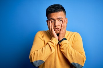 Young handsome latin man wearing yellow casual sweater over isolated blue background Tired hands covering face, depression and sadness, upset and irritated for problem