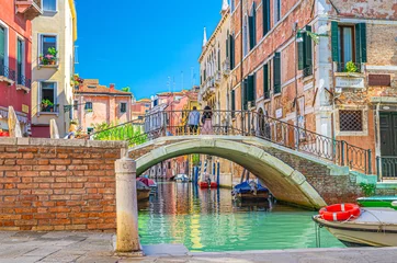 Foto op Canvas Brug over smal waterkanaal in Venetië met afgemeerde boten tussen oude kleurrijke gebouwen met balkons en bakstenen muren, blauwe lucht, regio Veneto, Noord-Italië. Typisch Venetiaans stadsbeeld © Aliaksandr