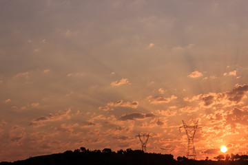 Mountain silhouette at sunrise. Sun rising between clouds on sky at morning. Pylons and trees on the mountain.