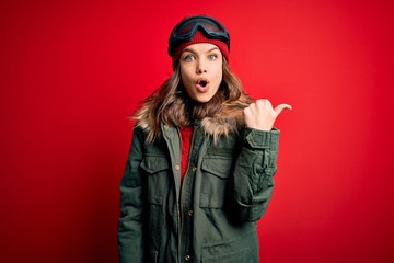 Young blonde girl wearing ski glasses and winter coat for ski weather over red background Surprised pointing with hand finger to the side, open mouth amazed expression.