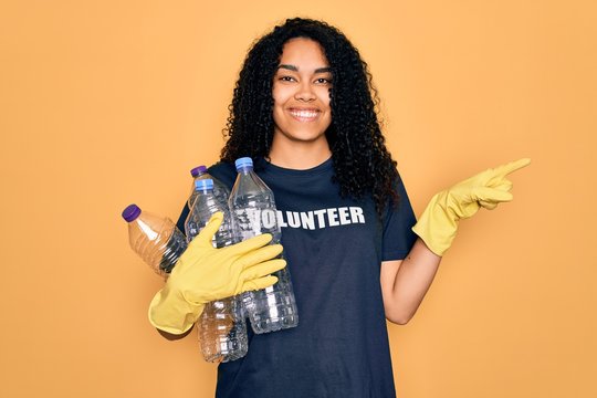 Young african american woman doing volunteering recycling holding plastic bottles very happy pointing with hand and finger to the side