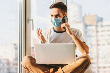 Young businessman talking on smart phone while sitting with laptop on laps on windowsill. Student freelancer having conversation on cell phone while working from home. Distance job with computer