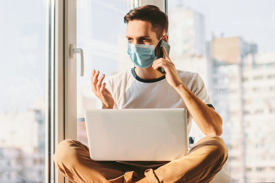 Young businessman talking on smart phone while sitting with laptop on laps on windowsill. Student freelancer having conversation on cell phone while working from home. Distance job with computer