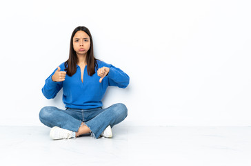 Young mixed race woman sitting on the floor isolated on white background making good-bad sign. Undecided between yes or not