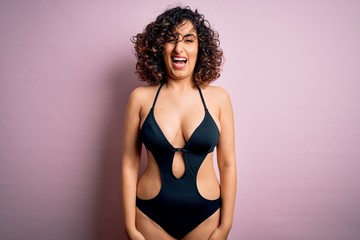 Young beautiful arab woman on vacation wearing swimsuit and sunglasses over pink background winking...