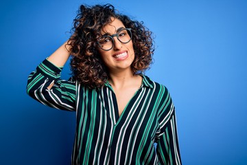 Young beautiful curly arab woman wearing striped shirt and glasses over blue background confuse and wonder about question. Uncertain with doubt, thinking with hand on head. Pensive concept.