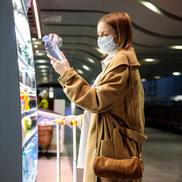 Caucasian woman customer wearing facial protective mask in airport, buying and choosing water bottle, reads information on a label. Purchase of food during the coronavirus, covid-19 epidemic in USA. 
