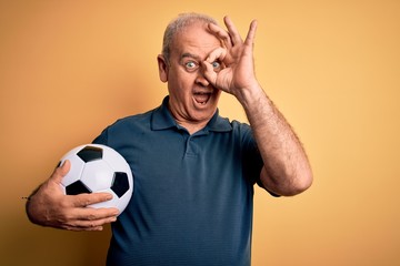 Middle age hoary player man playing soccer holding football ball over yellow background with happy face smiling doing ok sign with hand on eye looking through fingers