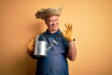 Middle age farmer man wearing apron and hat holding watering can over yellow background doing ok sign with fingers, excellent symbol