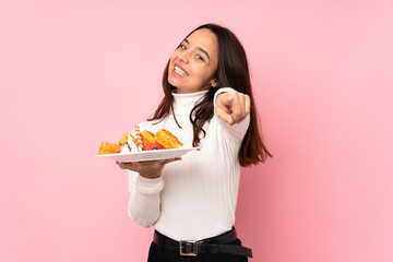 Young brunette woman holding waffles over isolated pink background making phone gesture and pointing front