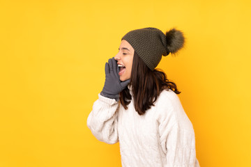 Young woman with winter hat over isolated yellow background shouting with mouth wide open to the lateral