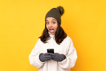 Young woman with winter hat over isolated yellow background surprised and sending a message