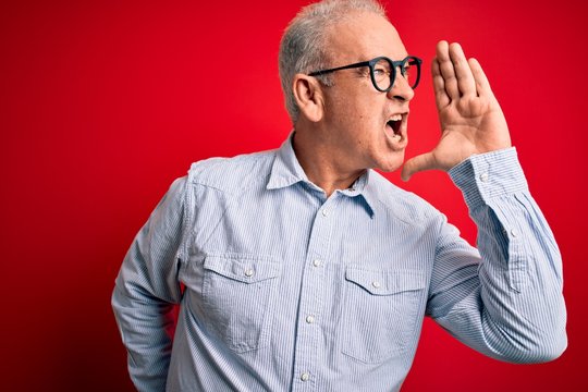 Middle age handsome hoary man wearing casual striped shirt and glasses over red background shouting and screaming loud to side with hand on mouth. Communication concept.
