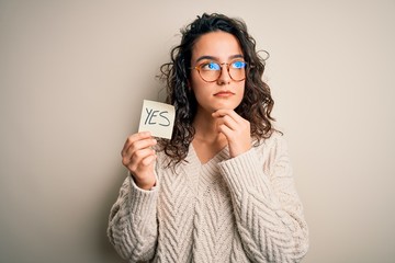 Young beautiful woman with curly hair holding reminder paper with yes word message serious face...