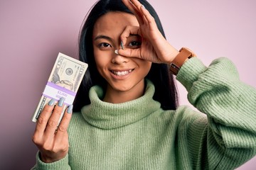 Young beautiful chinese woman holding dollars standing over isolated pink background with happy face smiling doing ok sign with hand on eye looking through fingers