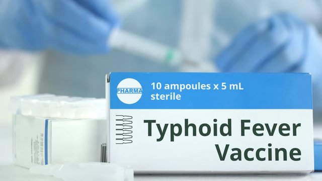 Box with typhoid fever vaccine on the table against blurred lab assistant or doctor. Fictional phaceutical logo