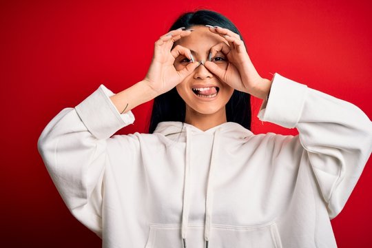 Young beautiful chinese sporty woman wearing sweatshirt over isolated red background doing ok gesture like binoculars sticking tongue out, eyes looking through fingers. Crazy expression.
