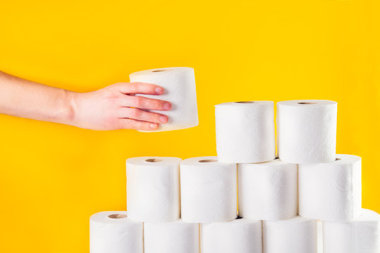 Close Up Caucasian Male Hand Takes A Roll Of Toilet Paper From A Folded Tower Of Rolls On Bright Yellow Background. Crisis Product Lack Concept.