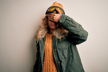 Young african american skier woman with curly hair wearing snow sportswear and ski goggles covering eyes with hand, looking serious and sad. Sightless, hiding and rejection concept