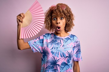 Young african american curly woman on vacation using hand fan over isolated pink background scared in shock with a surprise face, afraid and excited with fear expression
