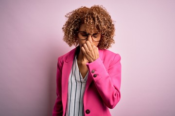 Young african american businesswoman wearing glasses standing over pink background smelling...