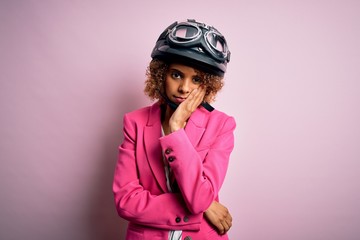 African american motorcyclist woman with curly hair wearing moto helmet over pink background thinking looking tired and bored with depression problems with crossed arms.