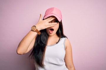 Young brunette woman wearing casual sport cap over pink background peeking in shock covering face and eyes with hand, looking through fingers with embarrassed expression.