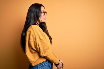 Young brunette woman wearing glasses and casual sweater over yellow isolated background looking to side, relax profile pose with natural face and confident smile.